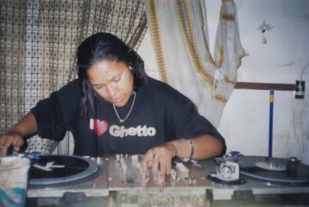 Image: Horizontal photo of Kay behind two turntables, one mixer, blending. Kay is looking down at the mixer, unaware of the photo being taken. Behind Kay are two mismatching curtains with two different design patterns—one is beige, elaborate tiling detailing while the other is primarily white with gold stripes; the walls are white. Kay’s hair is black, straight, shoulder-length. Kay is wearing a silver chain and a black T-shirt that says “ I (heart) Ghetto,” the heart is red, the text on the shirt is white. Photo courtesy of Kay. 