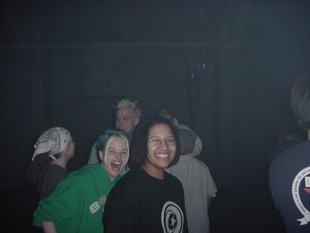 Image: Horizontal photo of Kay smiling wearing a black T-shirt with a white logo. Behind Kay is a white person wearing a green hoodie with a white logo. This person has blonde bangs which are longer than the rest of their hair that is blue. This person is photographed with their mouth open, smiling. Behind them are other white ravers, unaware of the photo being taken. One person is wearing a white bandana while the other has spiked, bleach blonde hair. The photo is taken indoors and with a flash. Photo courtesy of Kay.