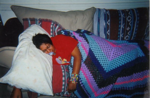 Image: Horizontal photograph of Kay asleep on a beige couch with a pillow without a pillowcase, a geometric lilac, periwinkle, cobalt, black and pink quilt. Kay is wearing a red short-sleeve T-shirt, eyeglasses, and various candy bead rave bracelets in different neon colors like green, blue, red, purple, white, and orange. Kay is sleeping with a pacifier in their mouth. The right couch pillow has a southwestern print throw on top. Image courtesy of Kay.