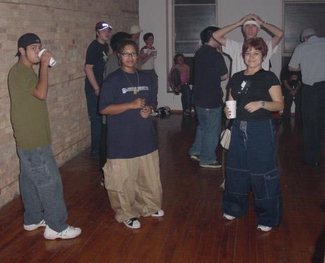 Image: Horizontal photo of Kay and party go-ers inside a dance space. To the left of Kay is a man wearing a black backward baseball cap, an army green colored T-shirt, blue cargo pants, and white sneakers. He’s the closest to a brick wall and looking at the camera while holding a white cup to his mouth. Next, is Kay wearing a navy T-shirt with a white logo, baggy khaki-colored JNCO pants, Adidas shell-toe sneakers, eyeglasses, and a chain. Kay’s has bangs that are longer than the rest of their hair. Kay is looking at the camera. Next to Kay is a woman wearing a black T-shirt with a white logo, blue baggy cargo pants, a beige crossbody bag, holding a white cup, smiling at the camera. Behind her, is a white man with a white T-shirt and appears to be looking at the camera. Behind him and next to him, are other people in the picture, a white wall with two windows where people are sitting on the ledge. The floor is a dark wood-paneled, contrasting the beige brick wall on the left and the white wall towards the back of the photo. Photo courtesy of Kay.