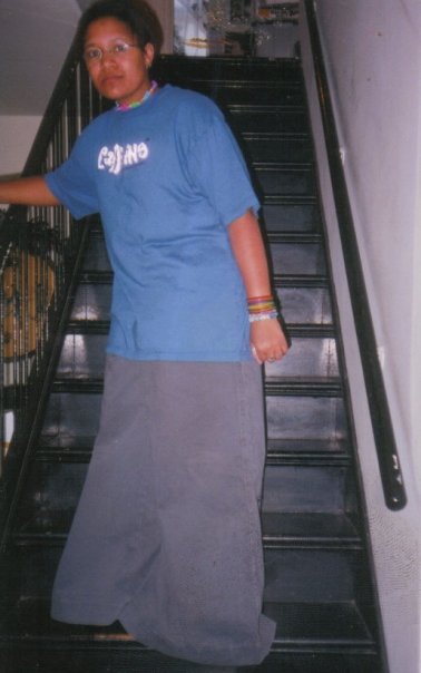 Image: Vertical photo Kay standing on a black staircase wearing eyeglasses, a blue short sleeve t-shirt that says “caffeine,” extremely baggy grey colored JNCO jeans, variously colored rubber and bead bracelets, a neon pink, green and blue choker. Their hair is short and out of their face. Photo courtesy of Kay.