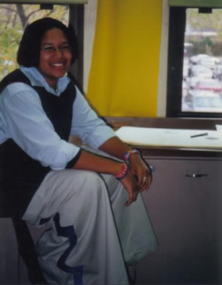 Image: Vertical photo of Kay sitting in what appears to be an art studio. Behind Kay are two windows, a yellow wall, a drawing pad with a pencil above a utility closet. Kay is smiling at the camera, wearing a silver chain, a blue button-down shirt with a black vest over it, baggy grey rave pants with a design on the side of the leg and a couple candy bead bracelets on each arm varying in colors like pink, light blue, orange and red. Courtesy of Kay.