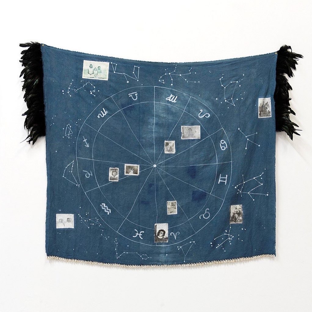 Image: Alexandria Eregbu, Cosmic Seed, 2018. Acrylic ink, cowrie shells, feathers, and indigo on linen. A multimedia fabric piece that is largely blue hangs on a white wall. White markings and black and white photographs cover the surface. Black feathers are on each side. Image courtesy of the artist.
