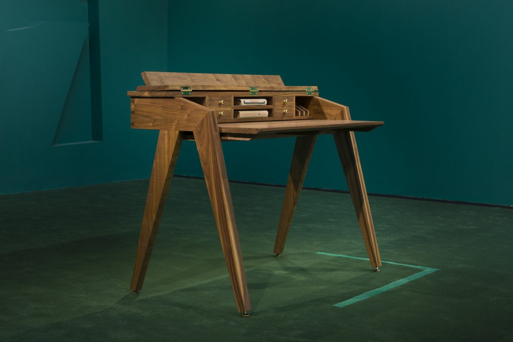 Image: An installation view of Caroline Kent's piece Writing Desk, 2021. Wood, 32h x 41w x 24d in, 81.28h x 104.14w x 60.96d cm. A wooden desk sits in the center of a green gallery. Image courtesy of the artist and PATRON Gallery, Chicago. Photography by Evan Jenkins.