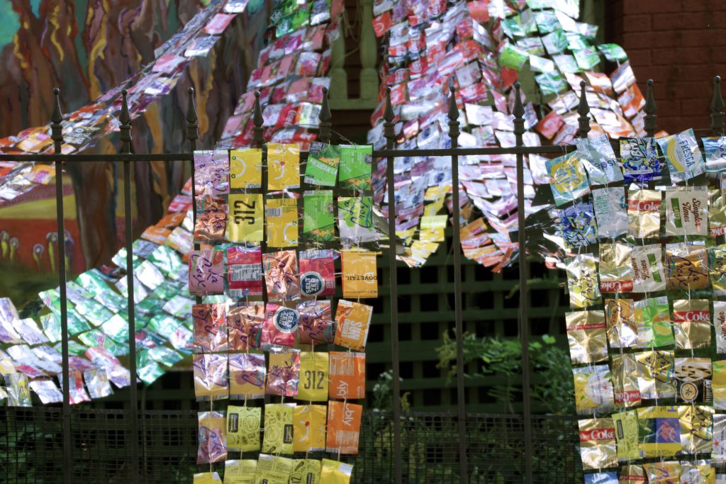 Image: Laurie LeBreton, “Collective Effervescence,” 2021. A close up look at 750 aluminum cans and 350 feet of waxed linen twine. Colorful ribbons patterned from cans are draped from the front porch and over the fence. Courtesy of Kitty Goldbert.