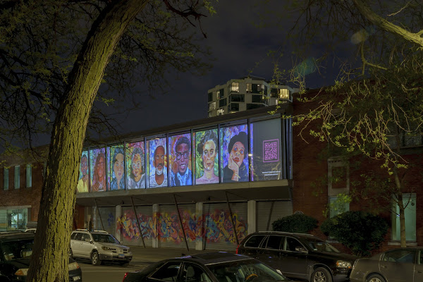 Image: A photo of the exterior of Hyde Park Arts Center at night. On the windows of HPAC are tiled projections of Zadov's colorful portraits, a bar of color in the dark. Behind HPAC is a building rising into the night sky. In front of it are a few cars lining the street and the trunk of a tree beginning at the bottom of the frame. Image courtesy of "What Time Is It?" Archive.
