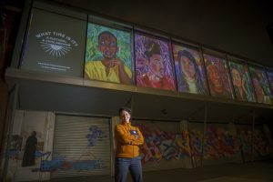 Featured Image: Irina Zadov standing outside of Hyde Park Arts Center at night. Above her, three colorful portraits are projected onto HPAC. Photo courtesy of the artist.