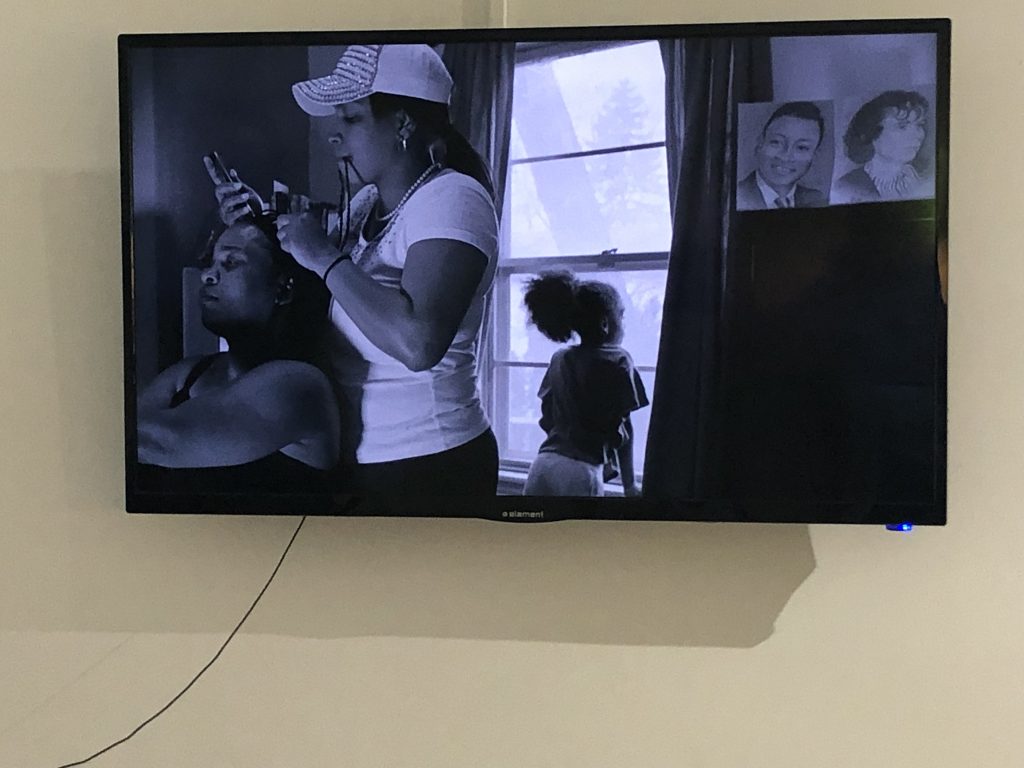 Image: Installation view of "Flint is Family." by LaToya Ruby Frazier, who captures intimate moments in the lives of residents in Flint, Mich. where they dealt with poisoned water. The image shows a still of the video, which is in blue-tinted black and white and includes a Black woman giving a haircut to a Black woman. A Black child looks out the window, and a vintage portrait of a black man and woman is on the wall behind the woman. Image courtesy of Bridgette Redman.