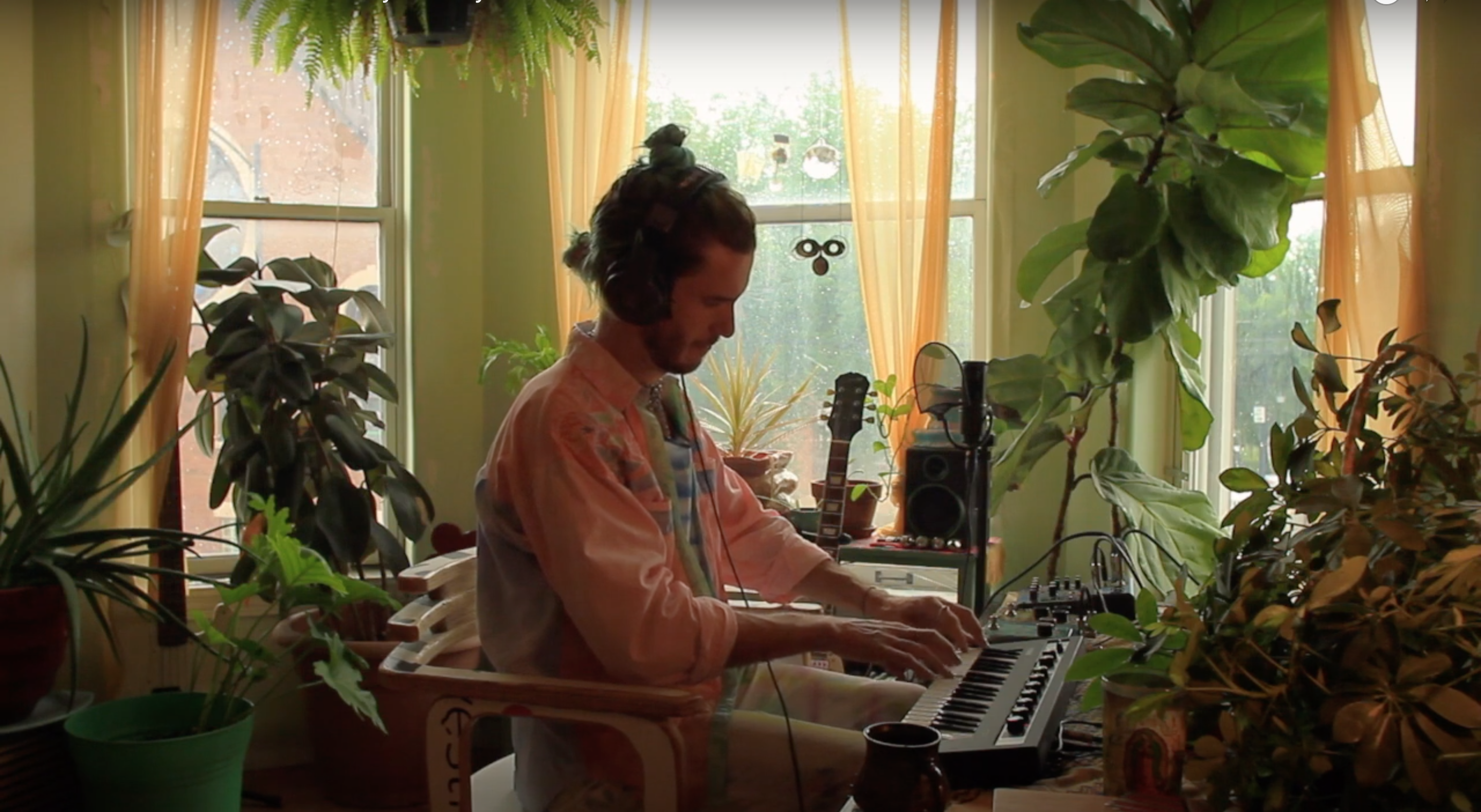 Jeffrey Michael Austin in their home studio performing A Place You Can Go. They are playing a keyboard and seated in front of three windows which are illuminating the room with natural light. The space around Jeffrey is filled with different types of houseplants.