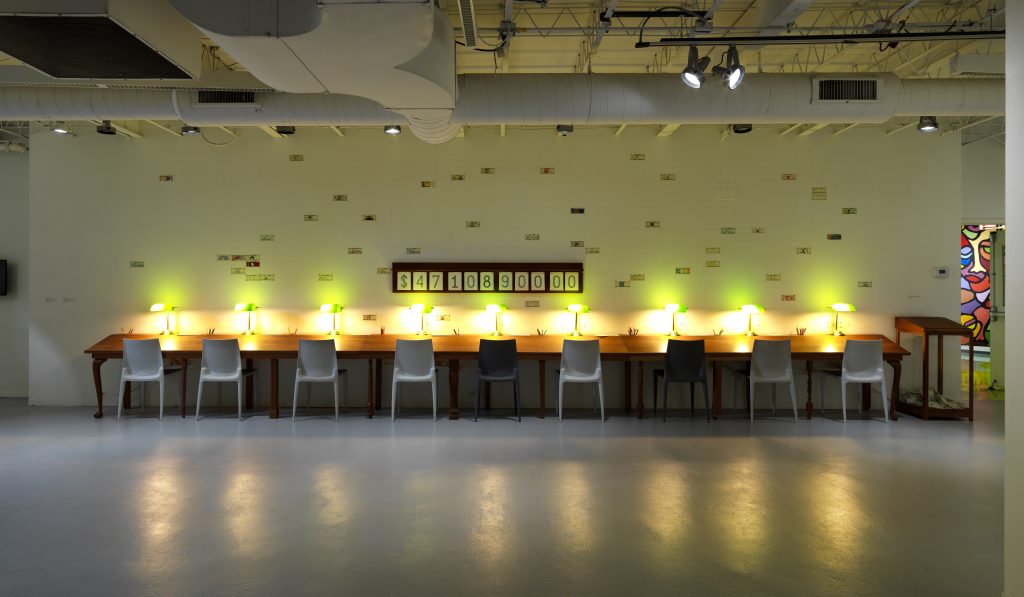 Image: Full installation view of Mel Chin's piece "Fundred Initiative: A bill for IL". The piece is comprised of a long table against a wall with nine green lamps sitting on the surface and nine chairs pulled up to the table. Many fake U.S. bills colored by children around the world are hung on the wall above the table. Image courtesy of Hyde Park Art Center.