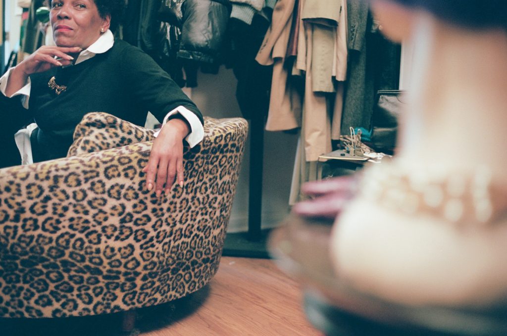 Image: A photo of Gilda Norris at Gilda Designer Thrift Boutique. She is sitting on a leopard-print chair wearing a black and white outfit. She wears tall platformed sneakers with stripes and a gold broach. All around her are various garments. She is looking to the right with one hand raised up to her chin. Photo by zakkiyyah najeebah dumas - o'neal.