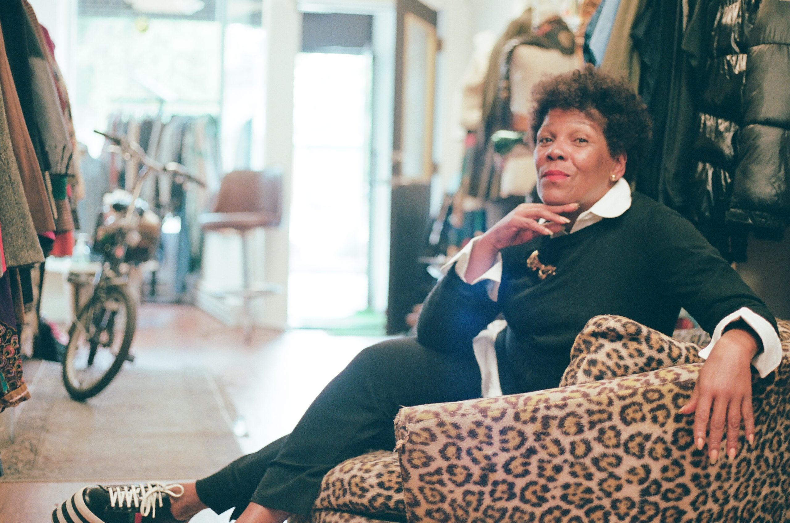 Featured image: A photo of Gilda Norris at Gilda Designer Thrift Boutique. She is sitting on a leopard-print chair wearing a black and white outfit. She wears tall platformed sneakers with stripes and a gold broach. All around her are various designer garments. She looks directly at the camera with one hand raised up to her chin. Photo by zakkiyyah najeebah dumas-o'neal.