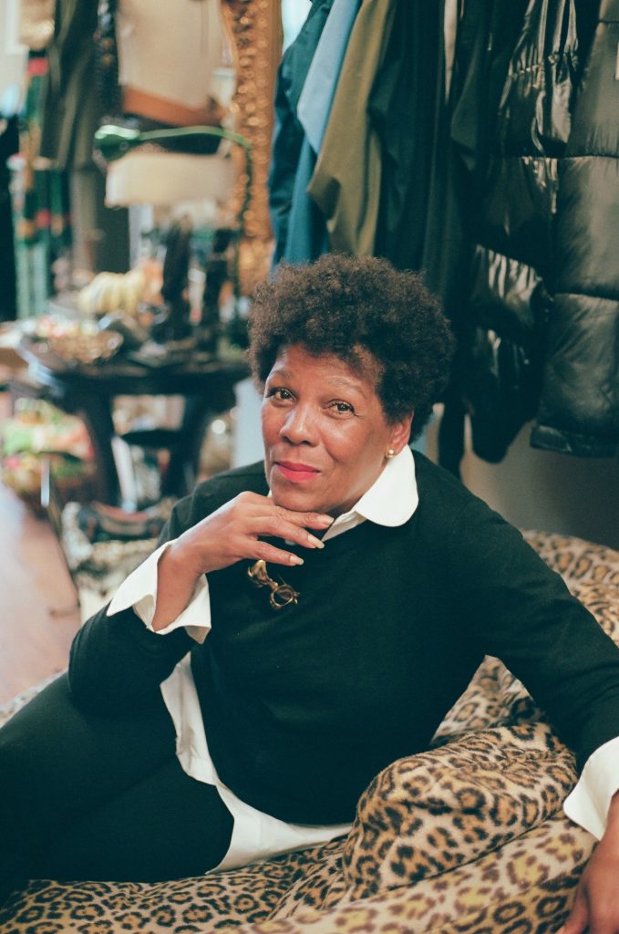 Image: A photo of Gilda Norris at Gilda Designer Thrift Boutique. She is sitting on a leopard-print chair wearing a black and white outfit. She wears tall platformed sneakers with stripes and a gold broach. All around her are various designer garments. She looks directly at the camera with one hand raised up to her chin. Photo by zakkiyyah najeebah dumas - o'neal.