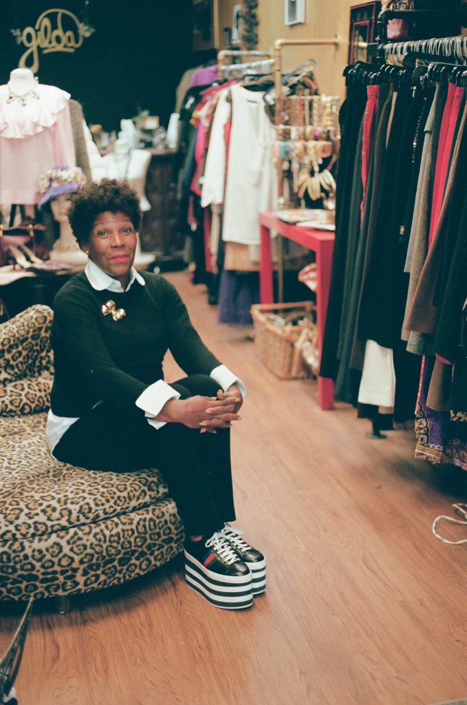 Image: A photo of Gilda Norris at Gilda Designer Thrift Boutique. She is sitting on a leopard-print chair wearing a black and white outfit. She wears tall platformed sneakers with stripes and a gold broach. All around her are various designer garments. She looks directly at the camera. Photo by zakkiyyah najeebah dumas - o'neal.