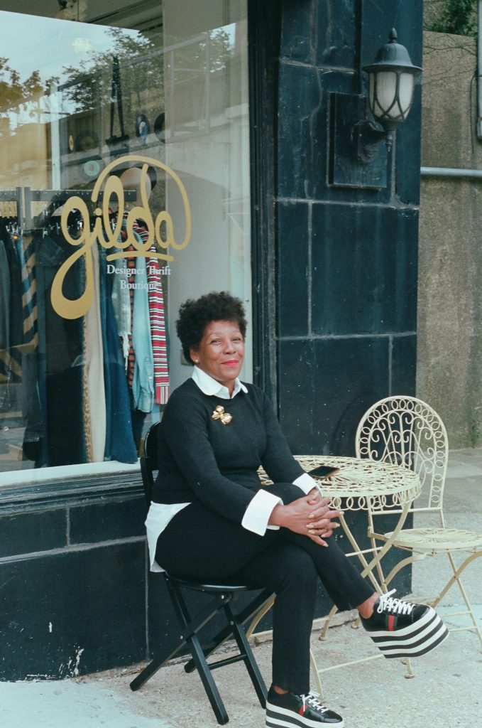 Image: A photo of Gilda Norris in front of Gilda Designer Thrift Boutique. She is sitting on a black chair in front of the store window, which says: "gilda" in looping gold letters. She is wearing a black and white outfit, tall platformed sneakers with stripes, and a gold broach. She looks directly at the camera with both hands resting on one knee. Photo by zakkiyyah najeebah dumas - o'neal.