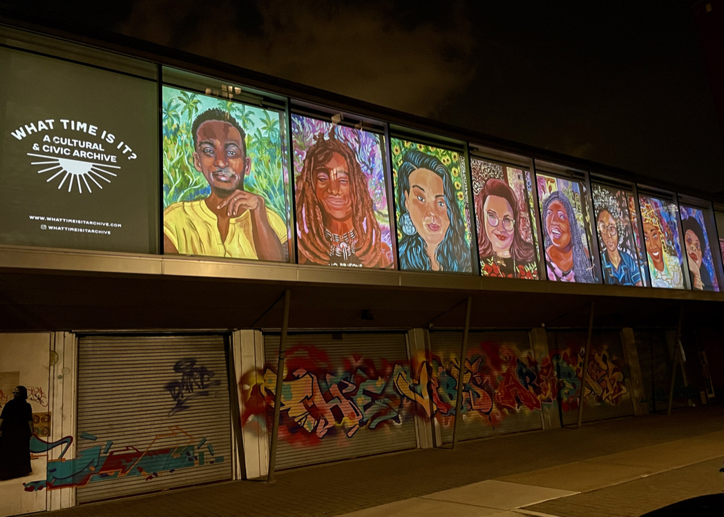 eatured Image: The exterior of Hyde Park Arts Center at night. Eight colorful portraits are projected onto HPAC. To the far left of the portraits is a dark square with the text: "What Time Is It? A Cultural & Civic Archive." Below the projected portraits are metal doors covered in red, blue, yellow, and black spray painted art. Photo courtesy of the artist.