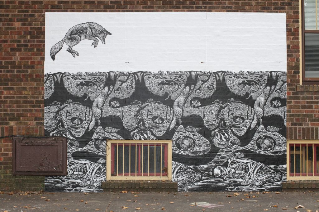 Image: Emmy Lingscheit, "Subterranea" at 717 N 5th Street, Springfield, IL. A black and white mural of a fox jumping in the air with tunnels being dug by rodents in the ground beneath. Photo by Jeff Robinson.