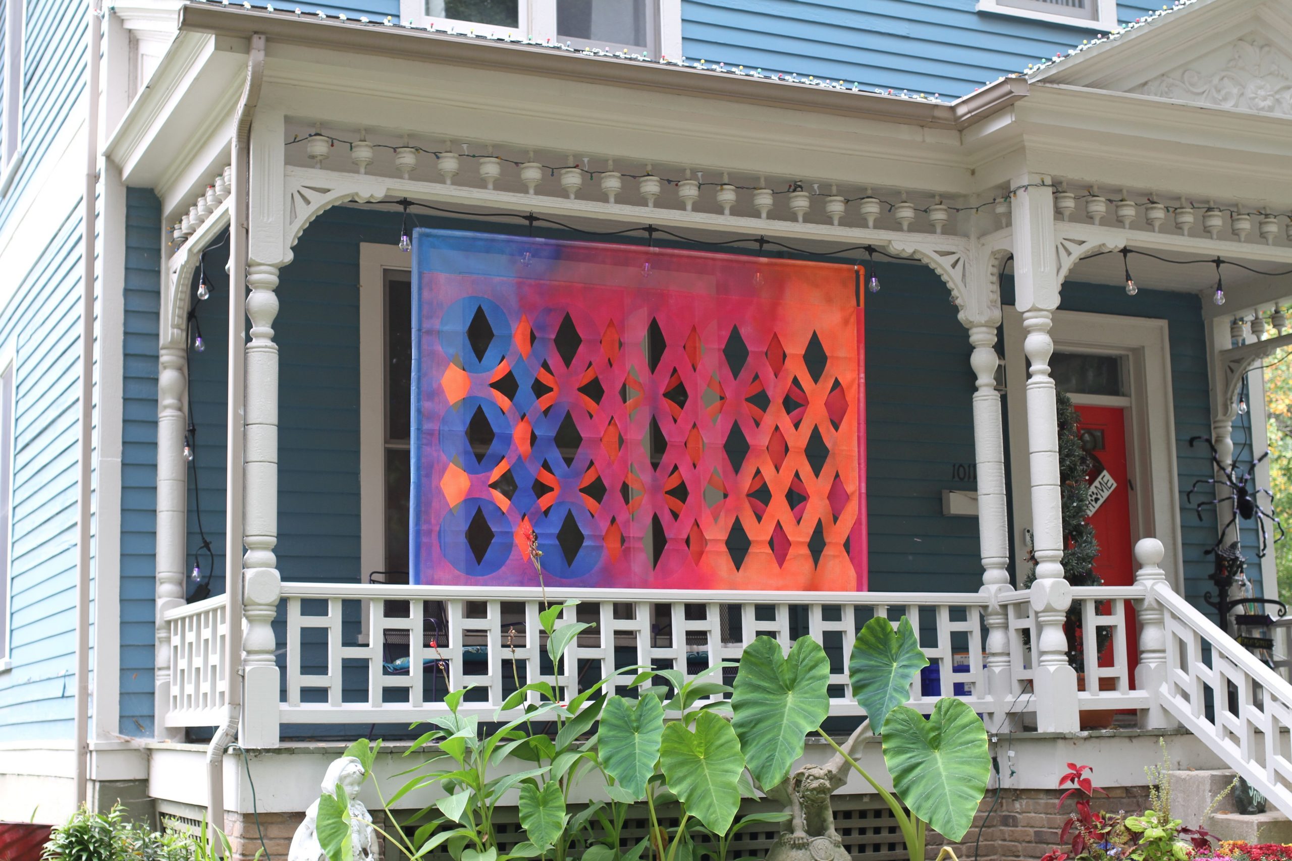 Featured image: Gina Hunt, XOXO at 1011 N. 6th Street, Springfield, IL. A colorful, geometric piece hangs in porch. The house is light blue with white trim. Plants grow in front of the porch. Photo by Jeff Robinson.