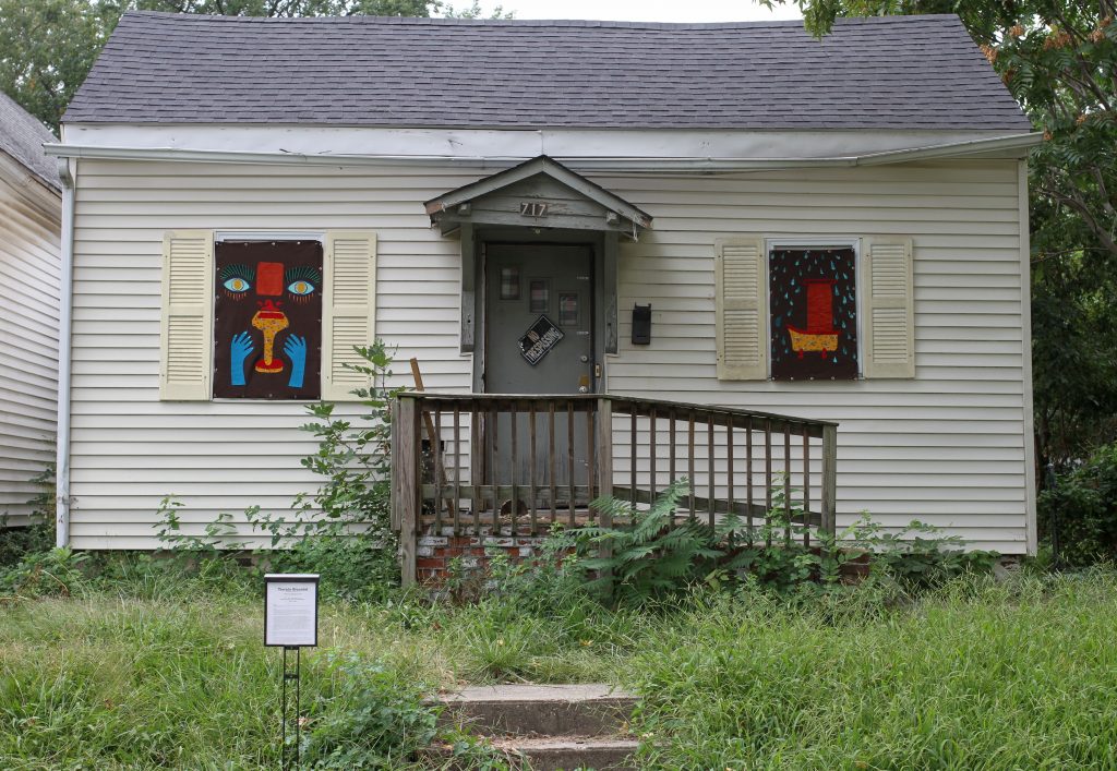 Image: Rachel Lebo and Janie Stamm, Take a peek at 717 N 7th Street, Springfield, IL. A white house with a grey door has artwork installed in front of its two windows. The artwork on the left has hands and a face, and the artwork on the right contains red and yellow shapes on a black background. Photo by Jeff Robinson.