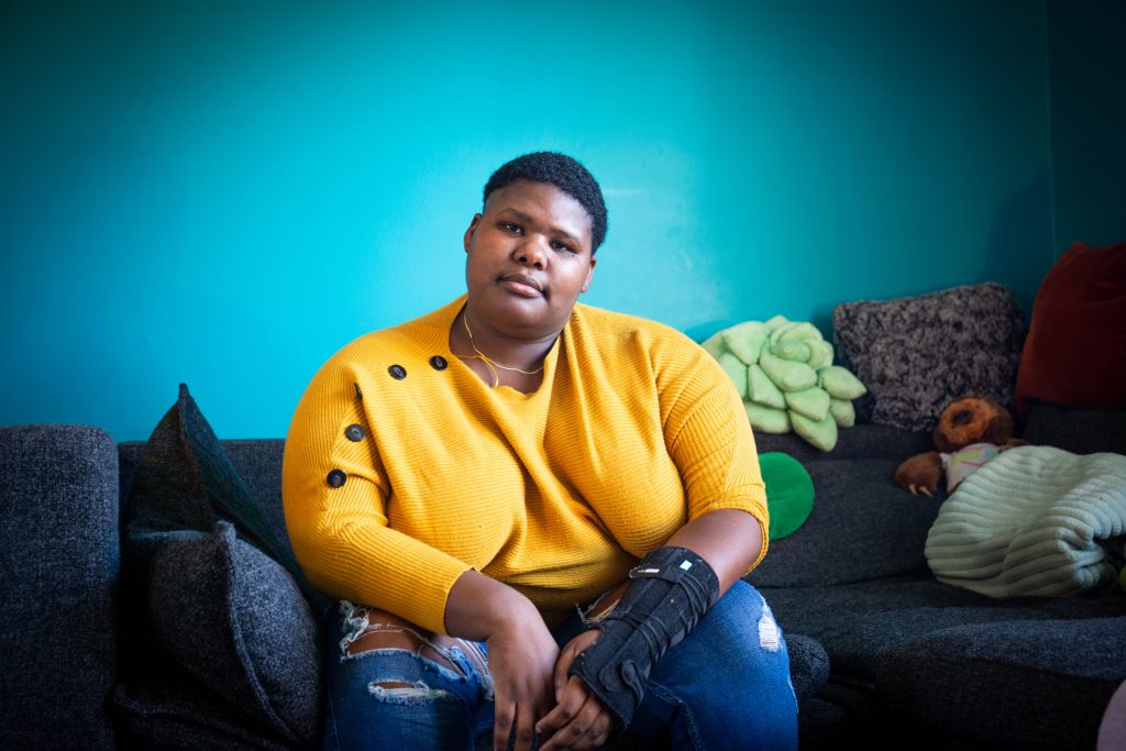 Image: A photo of Rise wearing a yellow sweater sitting on a grey couch in their home. They are looking directly at the camera. The wall behind them is teal. Photo by Ireashia Bennett.