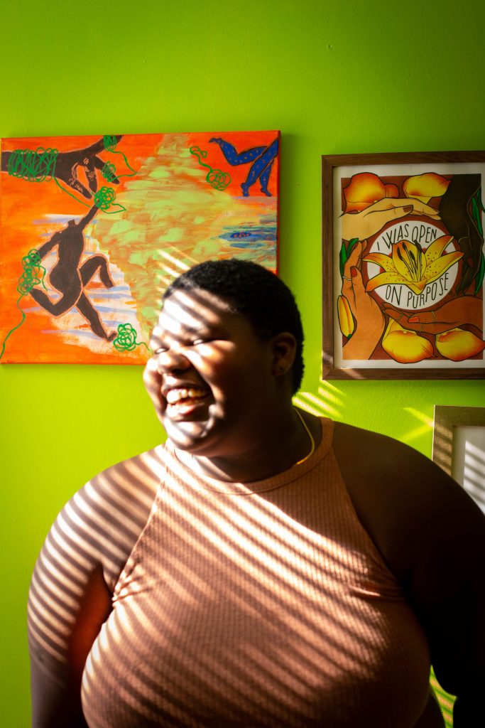 Image: A photo of Rise wearing a peach tank top while smiling. They are in front of a lime green wall in their home with different pieces of art hanging on the wall. Ribbons of light are coming from the left hand side of the frame and are shining on Rise, creating a striped pattern across their body. Photo by Ireashia Bennett.