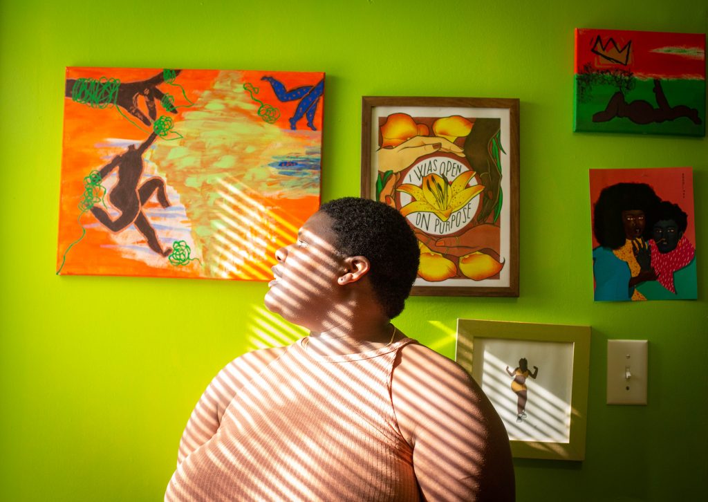 Image: A photo of Rise wearing a peach tank top while looking towards the light to our left. They are in front of a lime green wall in their home with different pieces of art hanging on the wall. Ribbons of light are coming from the left hand side of the frame and are shining on Rise, creating a striped pattern across their body. Photo by Ireashia Bennett.