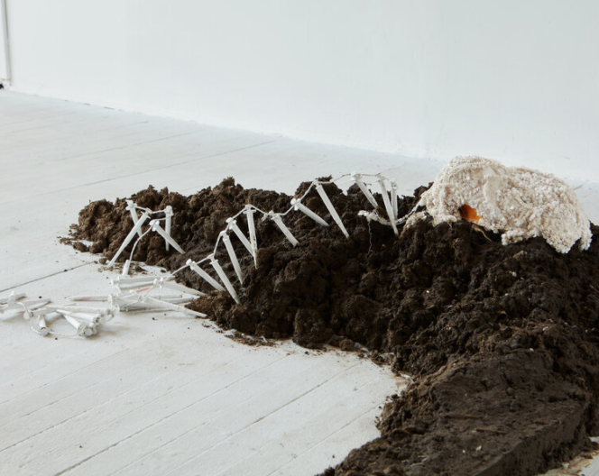 Image: A view of an installation piece by Sofia Moreno comprised of a mound of dirt with a sculpture of a dog on top and a row of tent pickets laid close by. Image by Ian Vecchiotti, courtesy of Hans Gallery.