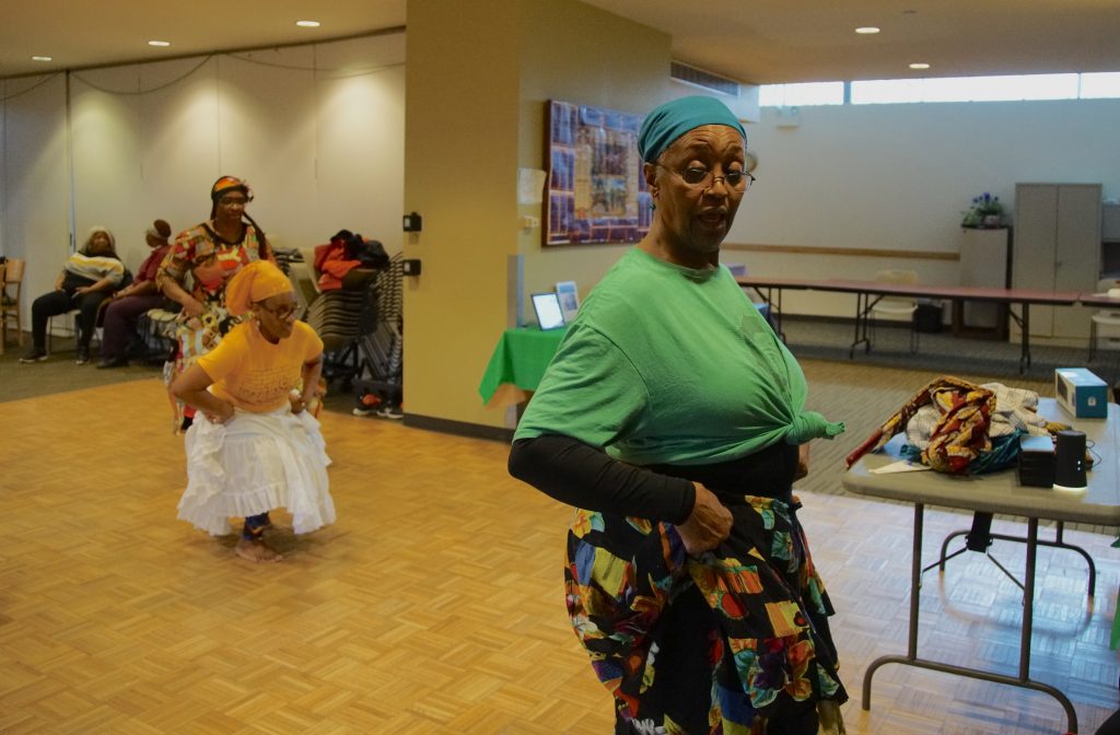 Image: Darlene Blackburn teaching at the Englewood Senior Center (2019). She is dancing while wearing a green shirt and a multicolored skirt. Photo by Wills Glasspiegel.