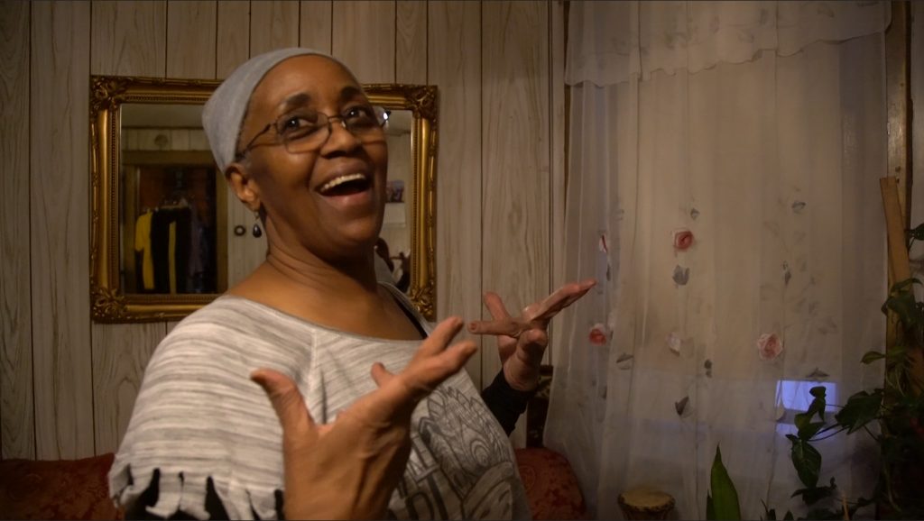 Image: Blackburn in her living room on the South Side of Chicago (2019). She is smiling wide while gesturing with both hands in the air. Photo by Wills Glasspiegel.