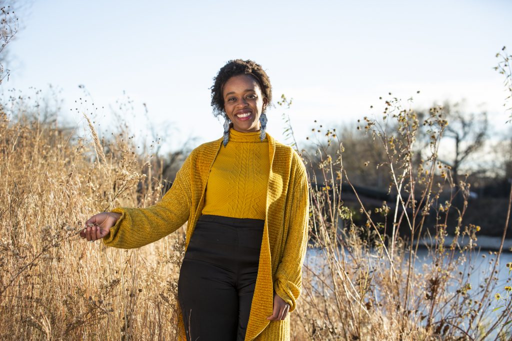 Image: Abena Motaboli stands in a wheat field in front of a lake wearing a yellow sweater and black pants. She smiles looking directly at the viewer with one hand outstretched touching the plants next to her. Photo by Kristie Kahns.