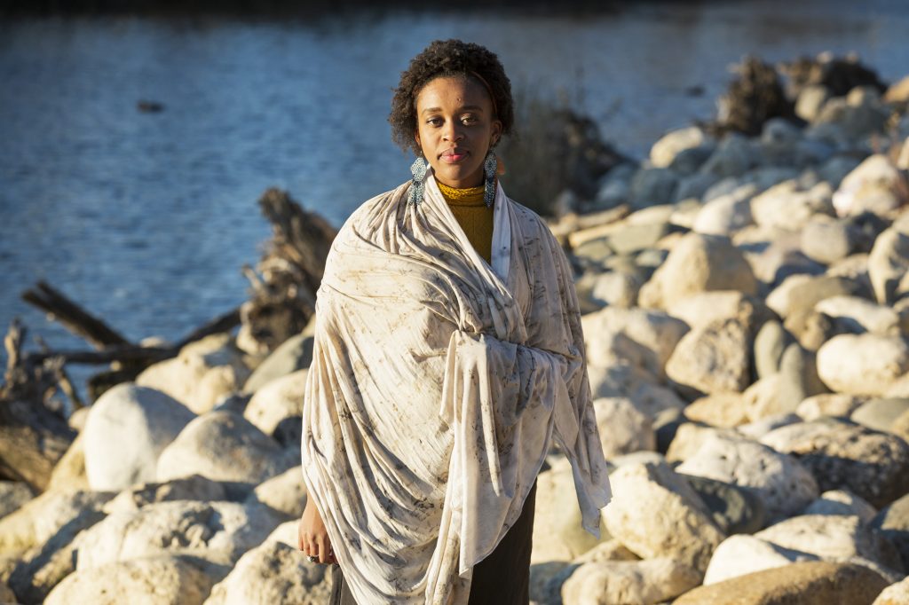 Image: Abena Motaboli stands in front of a lake and rocks wearing a yellow sweater and black pants. She is wrapped in a sheet of fabric that she has dyed with natural pigments. She looks directly at the viewer. Photo by Kristie Kahns.