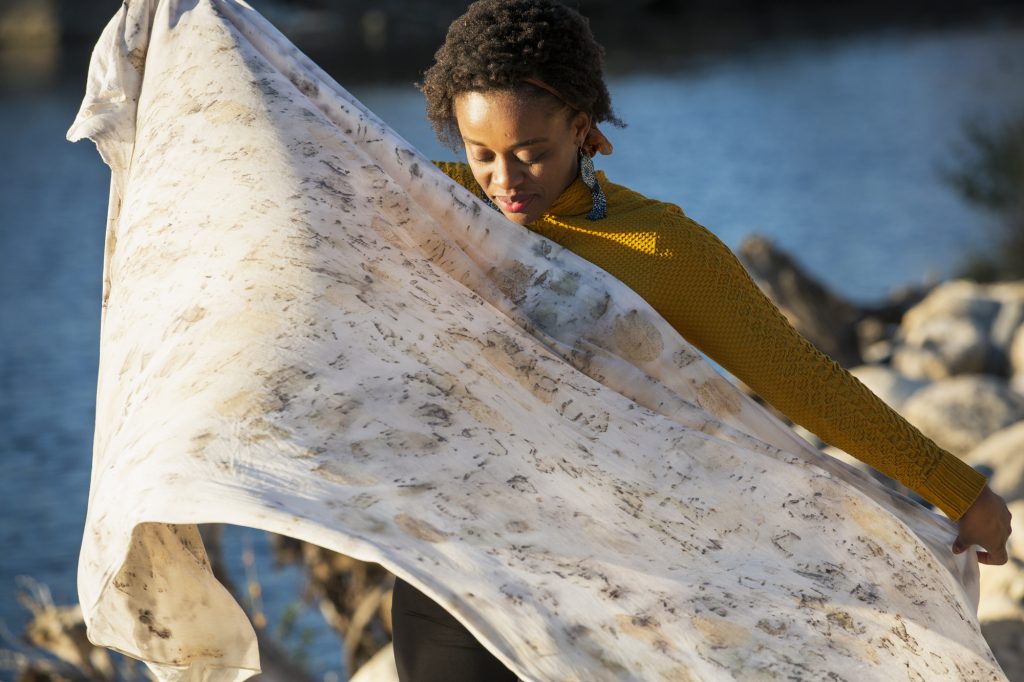 Image: Abena Motaboli stands in front of a lake wearing a yellow sweater and black pants. She holds up a sheet of fabric that she has dyed with natural pigments. Photo by Kristie Kahns.