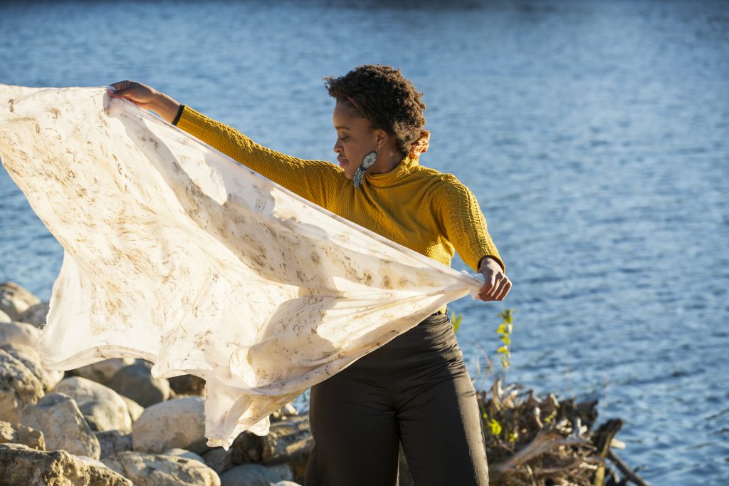 Image: Abena Motaboli stands in front of a lake wearing a yellow sweater and black pants. She holds up a sheet of fabric that she has dyed with natural pigments. Photo by Kristie Kahns.