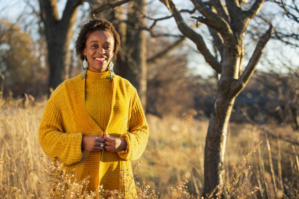 Image: Abena Motaboli stands in a wheat field near a tree wearing a yellow sweater and black pants. She smiles looking directly at the viewer. Photo by Kristie Kahns.