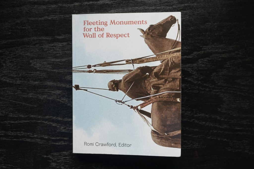 Image: A view of the cover of the book Fleeting Monuments for the Wall of Respect sitting on a black surface. The front cover has a sideways image of a public statue in the process of being taken down, with a partly cloudy blue sky in the background. The cover text reads "Fleeting Monuments for the Wall of Respect" in orange lettering and "Romi Crawford, Editor" in black lettering. The book's cover photo was provided courtesy of Valerie Cassel Oliver, 2020. Photo by Ryan Edmund Thiel.