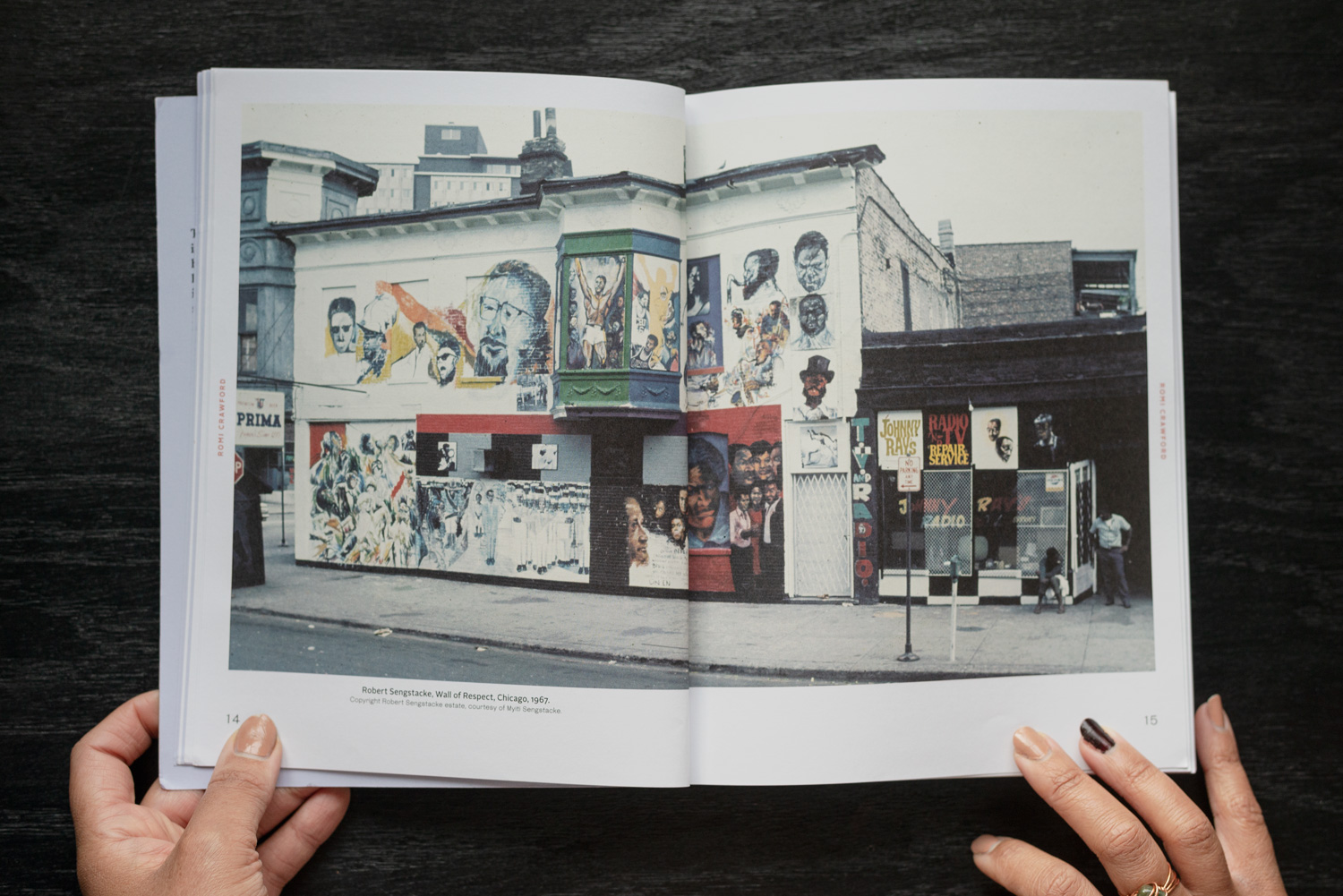 Image: The book Fleeting Monuments for the Wall of Respect is open to pages 14 and 15, showing a 1967 photo by Robert Sengstacke of The Wall of Respect in Chicago. You see a street-level view of a mural composed of over a dozen portraits of historic figures painted on the side of a two-story building. Photo by Ryan Edmund Thiel.