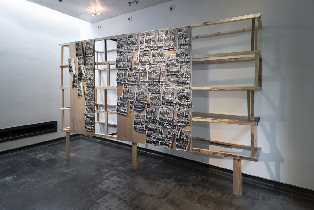 Image: Amina Ross, detail of Man’s Country installation at Iceberg Projects, 2021. A large wooden scaffold structure that is partially faced with plywood and weathered black and white Xeroxed flyers. Image courtesy of Iceberg Projects.