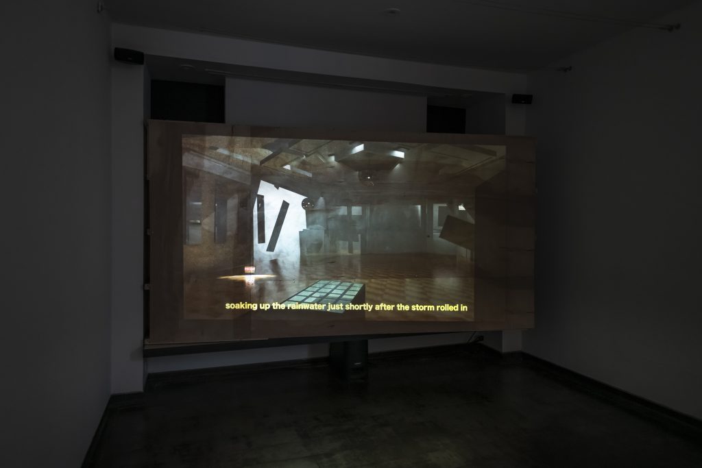 Image: Amina Ross, detail of Man’s Country installation at Iceberg Projects, 2021. A plywood faced screen pops out from the wall, more on the right side than on the left, and shows a still of a projected image of a digitally animating club with the subtitles soaking up the rainwater shortly after the storm rolled in. Image courtesy of Iceberg Projects.