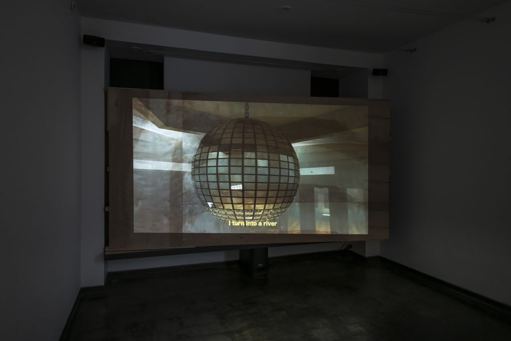 Image: Amina Ross, detail of Man’s Country installation at Iceberg Projects, 2021. A plywood faced screen pops out from the wall, more on the right side than on the left, and shows a still of a projected image of a digitally animating disco ball with the subtitles I turn into a river. Image courtesy of Iceberg Projects.