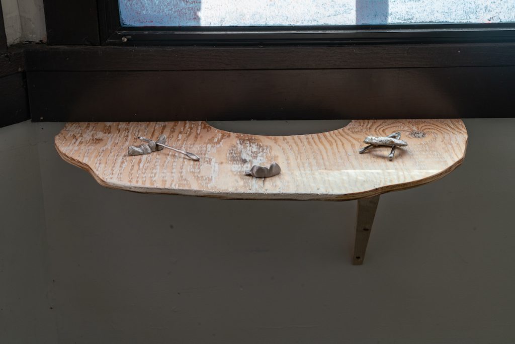 Image: Installation view of UTENSIL at Comfort Station. Three small pewter works led by Maggie Wong and created by Maggie Wong and Alden Burke are displayed on a curved, wooden shelf. Image courtesy of Annas.