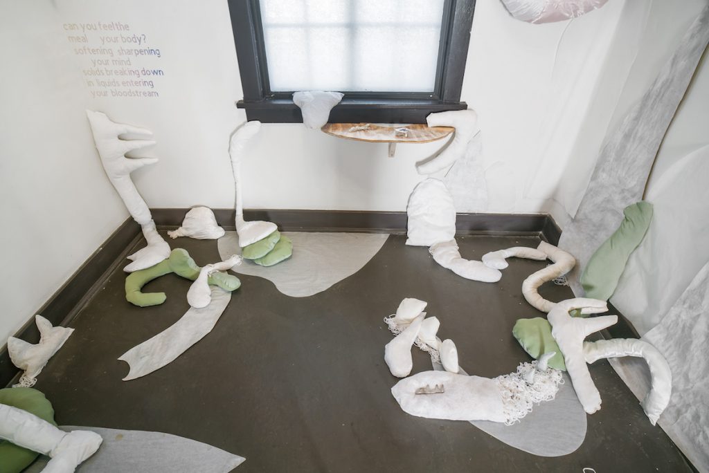 Image: Installation view of UTENSIL at Comfort Station. White, grey, and light green soft sculptures led by Mariel Harari and created by Mariel Harrari and Caroline Dahlbergare are displayed throughout the space. On the back wall, there is vinyl text on the left and a wooden shelf in the middle displaying small sculptural work by Maggie Wong. Image courtesy of Annas.