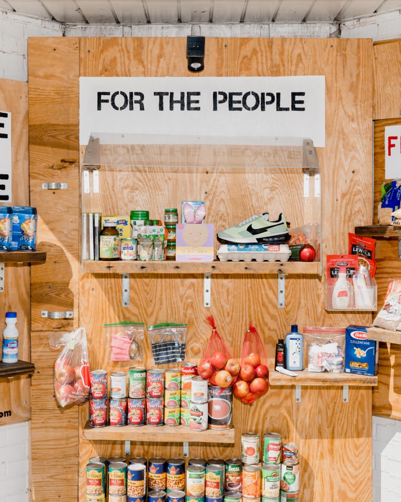 Image: “alt_ market: Greater Grand Crossing.” The Market serves as a functional art installation that transforms abandoned spaces into a communal free market to create a temporary communal shared economy. The image shows items available in alt_ market (including canned goods, bags of apples, baby food, masks, eggs, toiletries) in a plywood display. A sign on the installation reads, “For the people.”  Photo by Lyndon French. 