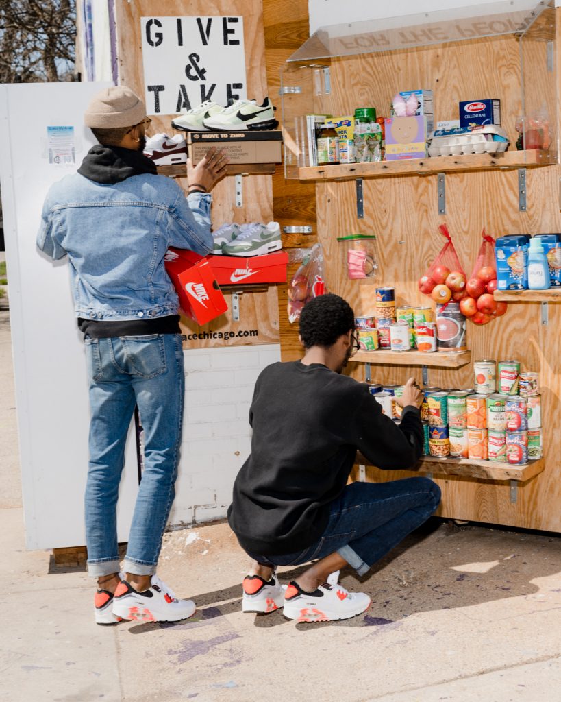 Image: Jordan Campbell and Jon Veal stock sneakers and canned goods in alt_ market installation. A sign on the installation reads, “ Give & take.”  Photo by Lyndon French. 