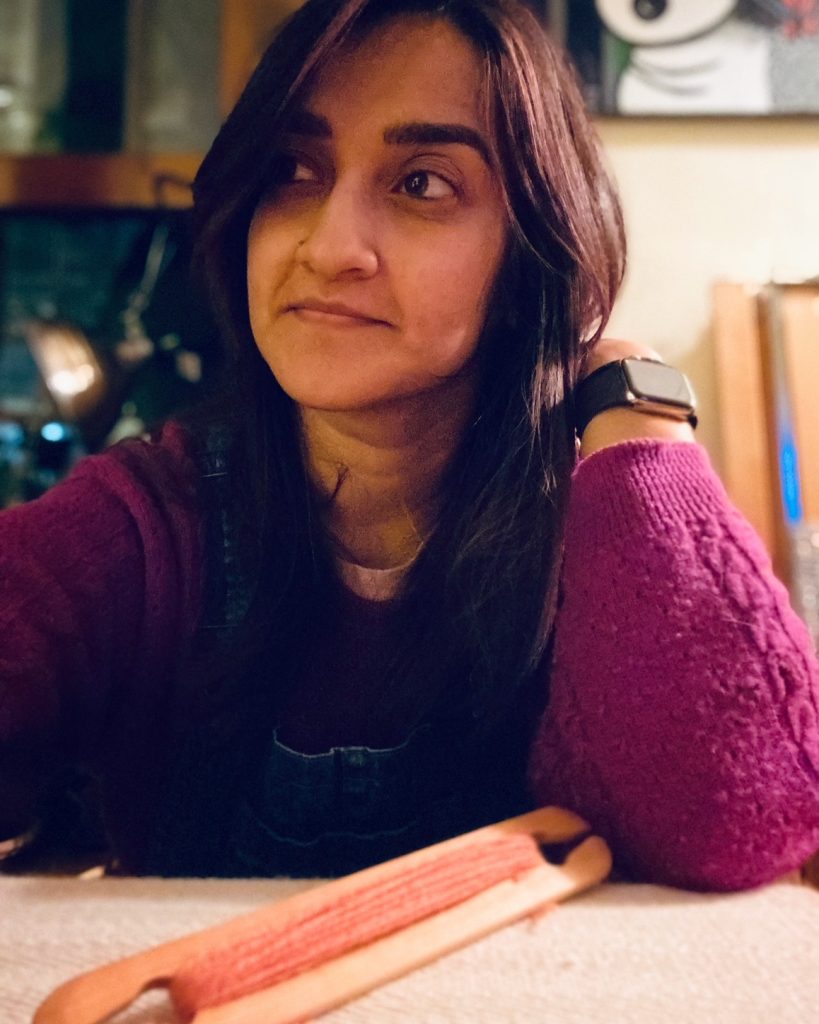 Image: A photo of Sangi Ravichandran. She sits at a table wearing a magenta sweater. She has one arm up on the table and looks slightly to the upper left corner of the composition. Photo courtesy of the artist.