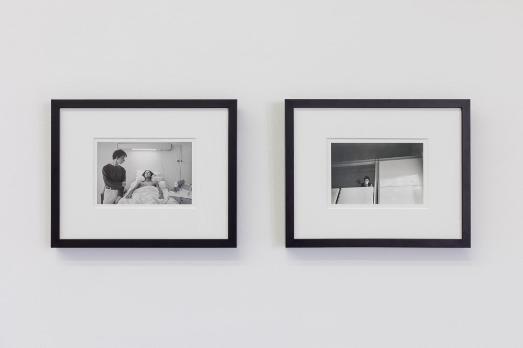 Image:Two framed black and white photos hang on a white wall. The image on the left is of two people, one standing the other in a hospital bed. The image on the right is a person looking down at the viewer from a balcony. Hervé Guibert, Les Amis, 1982 (left) AND C á la villa, N.D. (right). Photo: Useful Art Service.