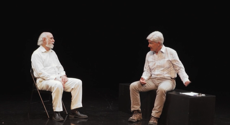 Image: Actors Bob Allen and Brian Robertson perform a scene as 'White Category' and the Patient respectively. They sit across form each other in a dark room; one in a chair, the other a bench. They both wear soft white and cream colored shirts and trousers. They are engaged in conversation. Image courtesy of Mourning the Creation of Racial Categories Project.