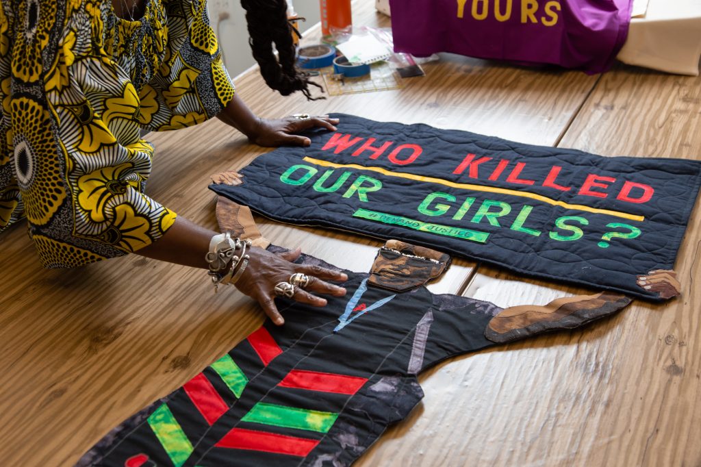 A focused photo of Dorothy Burge's hands and a portrait patch of a person holding a black sign that reads "WHO KILLED OUR GIRLS" in red, yellow, and green lettering. The protest portrait quilt is displayed on a table surface. Photo by Joshua Clay Johnson.