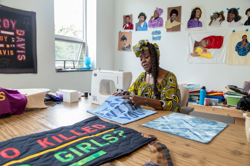 A portrait of artist Dorothy Burge sitting at a sewing machine in her studio at Hyde Park Art Center. In the foreground are various quilt pieces and sewing materials. Behind her are more sewing materials as well as artworks hanging on the walls. Photo by Joshua Clay Johnson.