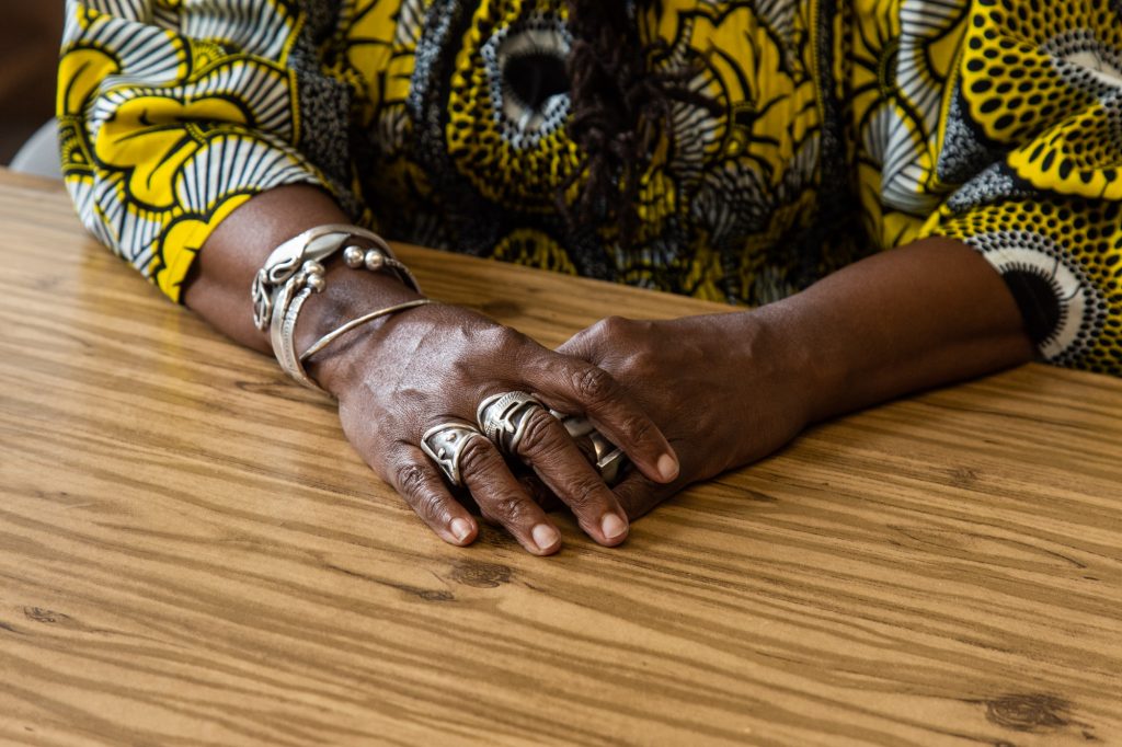 A focused photo of Dorothy Burge's hands, folded on a brown table. You catch a glimpse of her top's black, yellow, and white patterns. She is wearing silver bracelets and rings on multiple fingers. Photo by Joshua Clay Johnson.