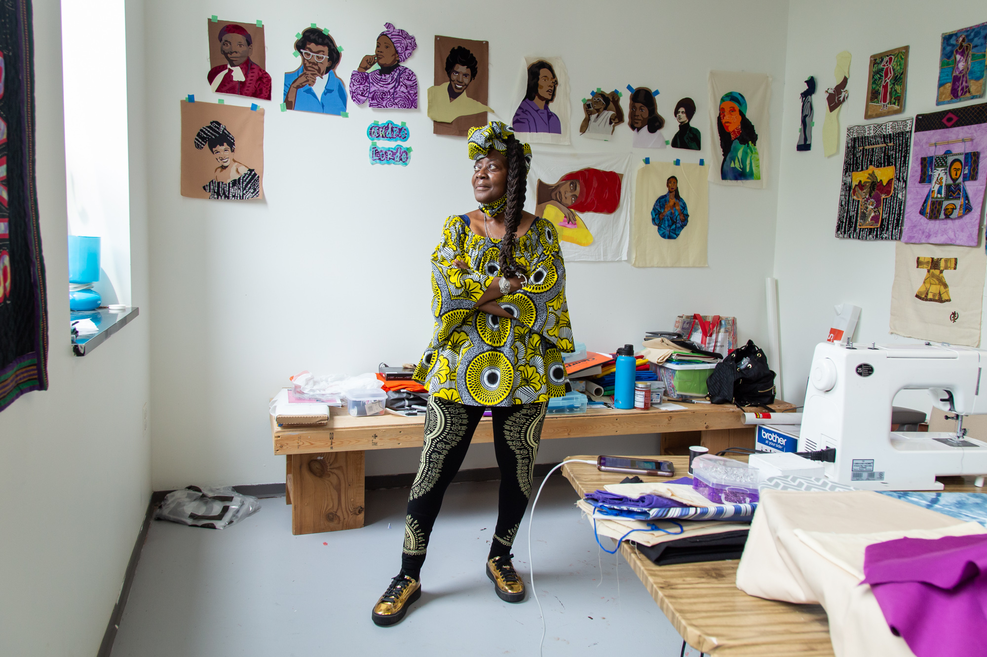 A full-length portrait of artist Dorothy Burge standing in her studio at Hyde Park Art Center. She wears a top, head wrap and pants with black, yellow, and white patterns. In the foreground is a table with a sewing machine and sewing materials. Behind her is another bench with sewing and other materials. Along the wall are fabric portraits of iconic Black women including Lena Horne, Harriet Tubman, Gwendolyn Brooks, Coretta Scott King, Kathleen Cleaver, and others. Photo by Joshua Clay Johnson.